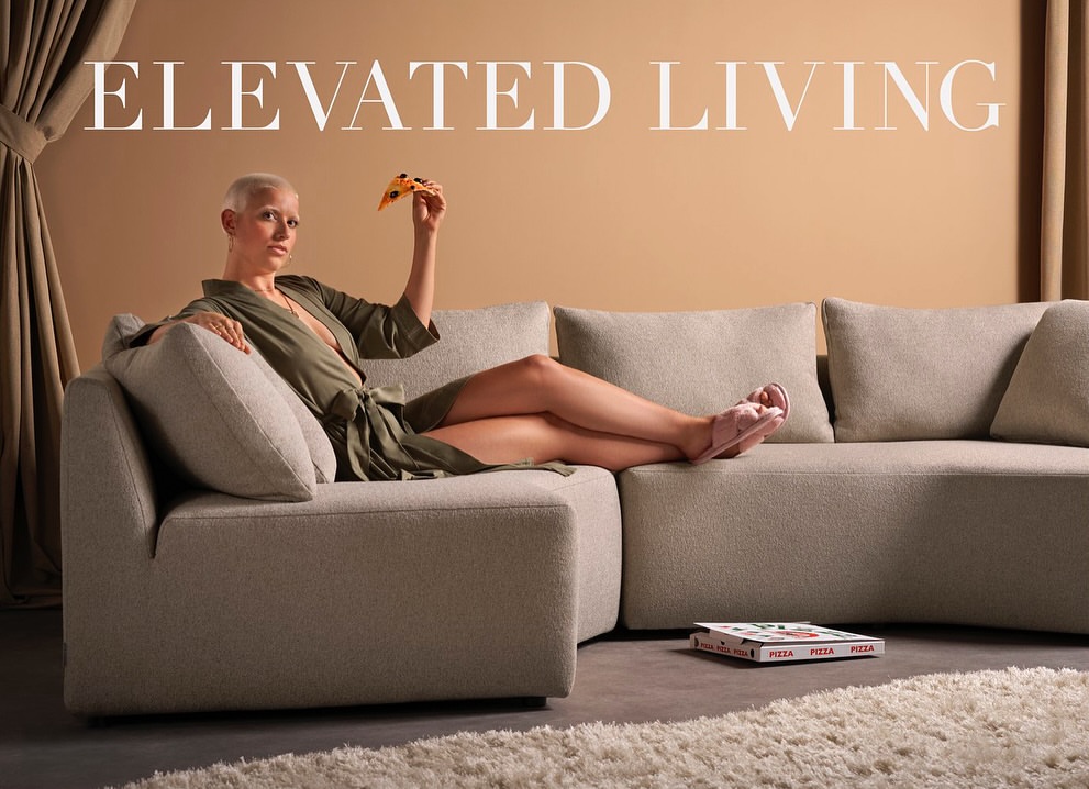 Woman in robe lounges on a modern sofa in a neutral interior, holding a slice of pizza; pizza box on floor.
