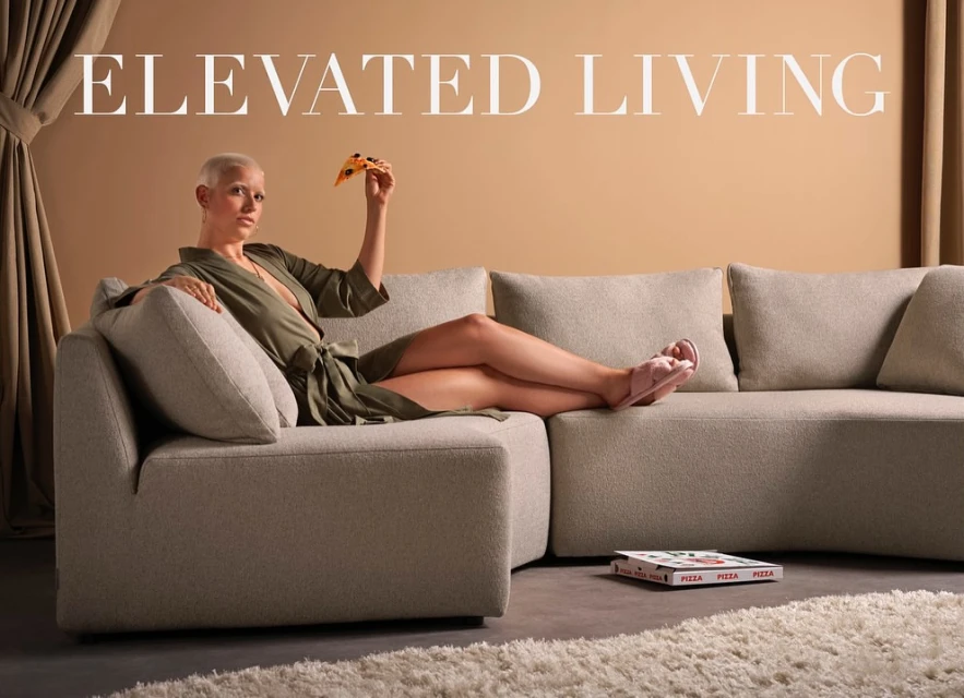 Woman in robe lounges on a modern sofa in a neutral interior, holding a slice of pizza; pizza box on floor.