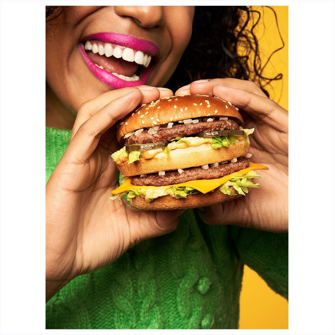 Close-up of an adult woman holding a large double cheeseburger with lettuce, pickles, onions, and special sauce on a yellow background.