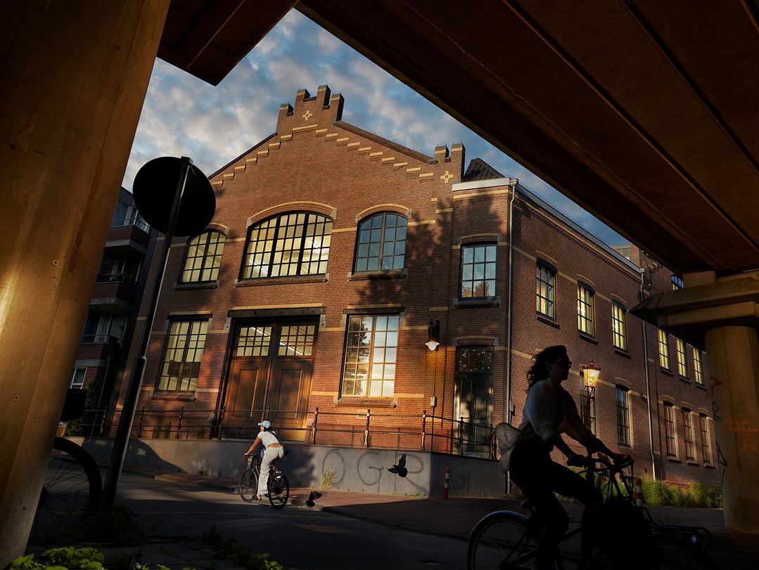 Two cyclists ride on the street in front of an old brick building under a bridge, bathed in warm sunlight during the day.