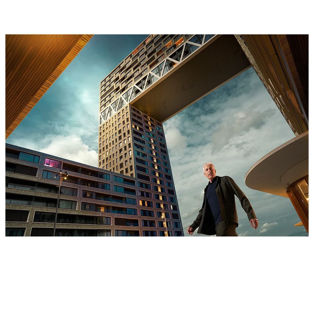 Adult male in dark jacket and pants walks near modern buildings under a cloudy sky, architecture featuring sharp lines and glass windows.
