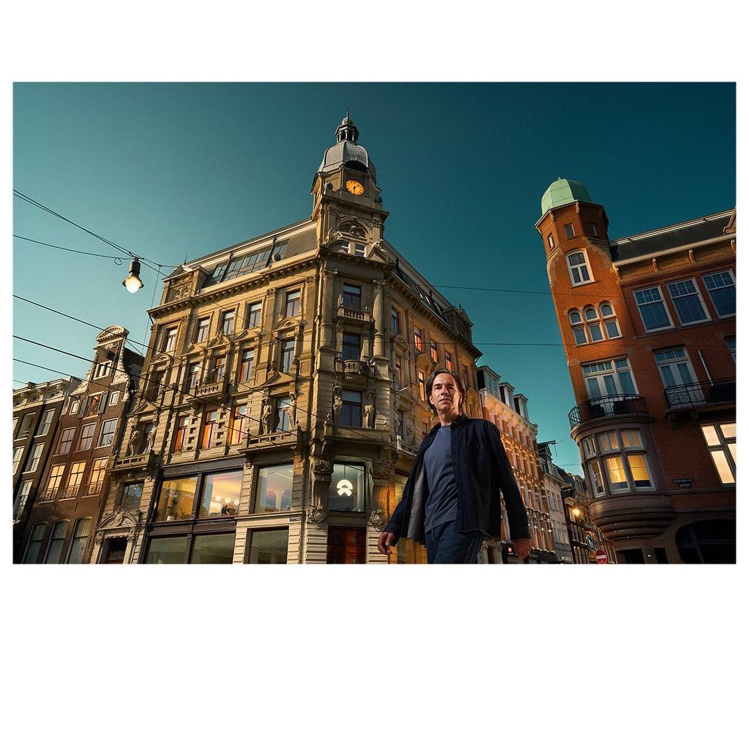 Adult male walks in Amsterdam, Netherlands, with historic buildings, clock tower, street lights, and teal sky as a backdrop.