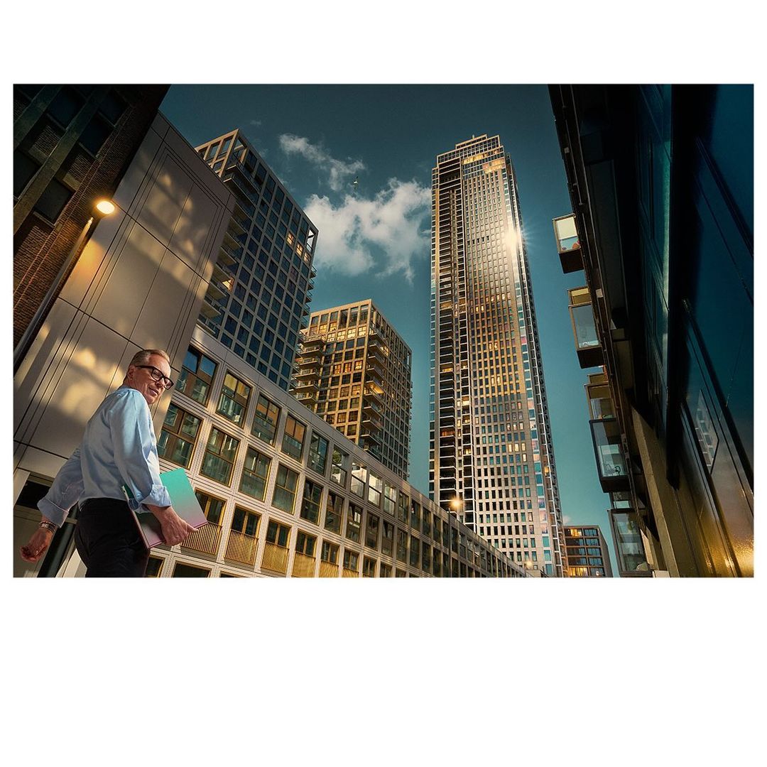 Low angle shot of a smiling man with glasses carrying a laptop in an urban city setting with tall buildings and a blue sky backdrop.