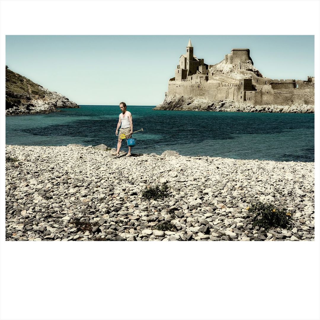 A man walks on a rocky beach with a bucket and shovel near the ocean, with an island castle in the background on a sunny day.