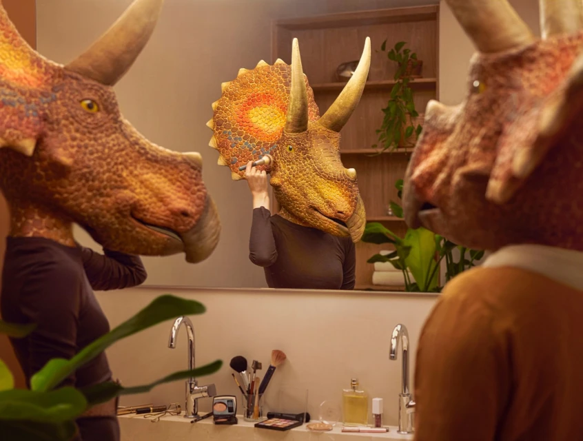 Triceratops-headed figures in a bathroom applying makeup reflected in a mirror, with bathroom sink and plant details.
