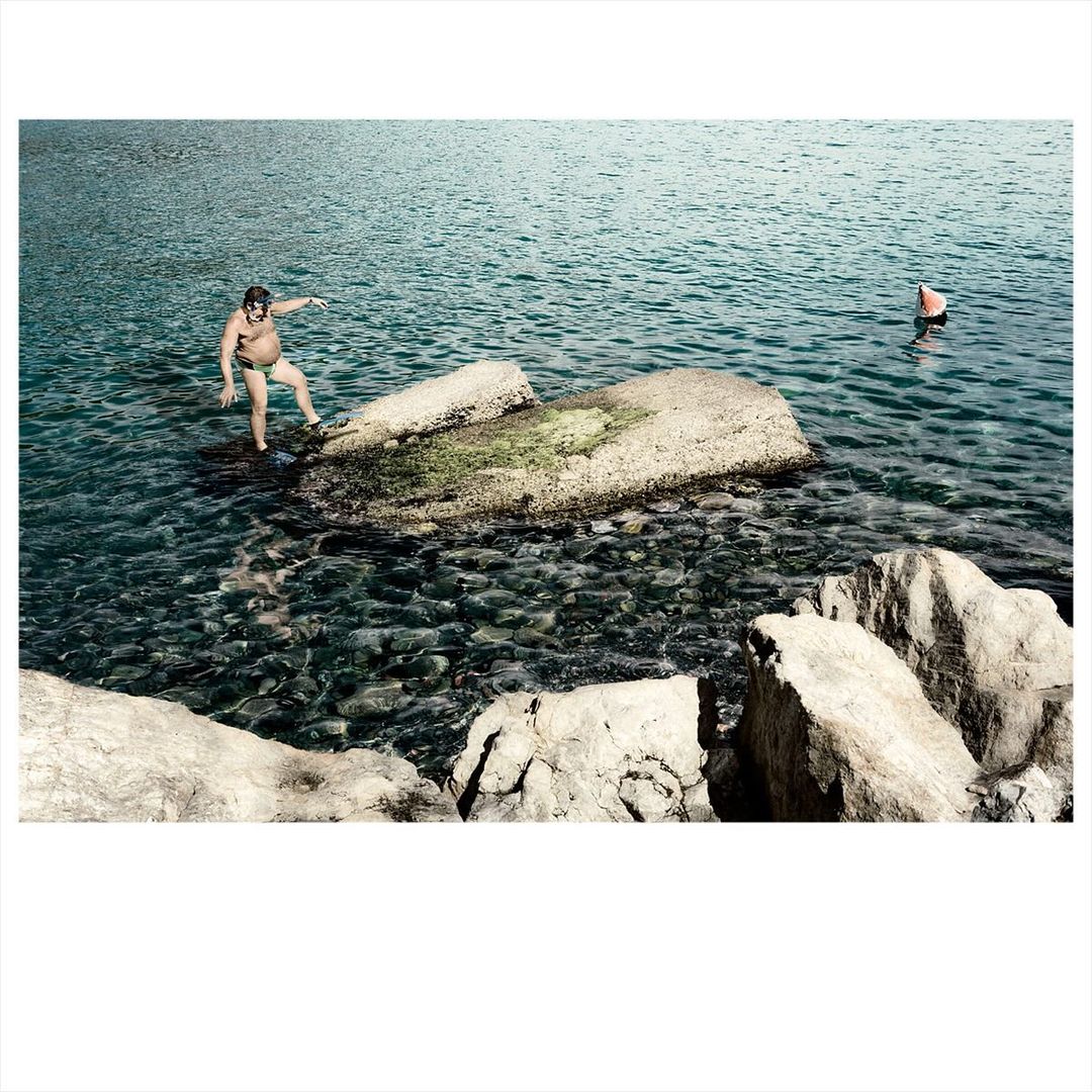 An adult male stands on rocks in clear water with a buoy in the background, captured in a medium shot with natural lighting.