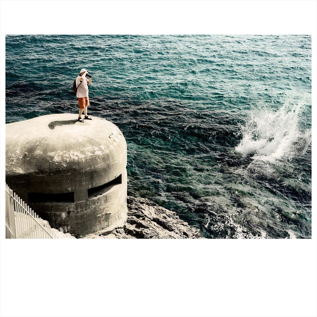 A man stands on a concrete bunker near the ocean, capturing a photograph of the splashing waves against the rocky coastline.