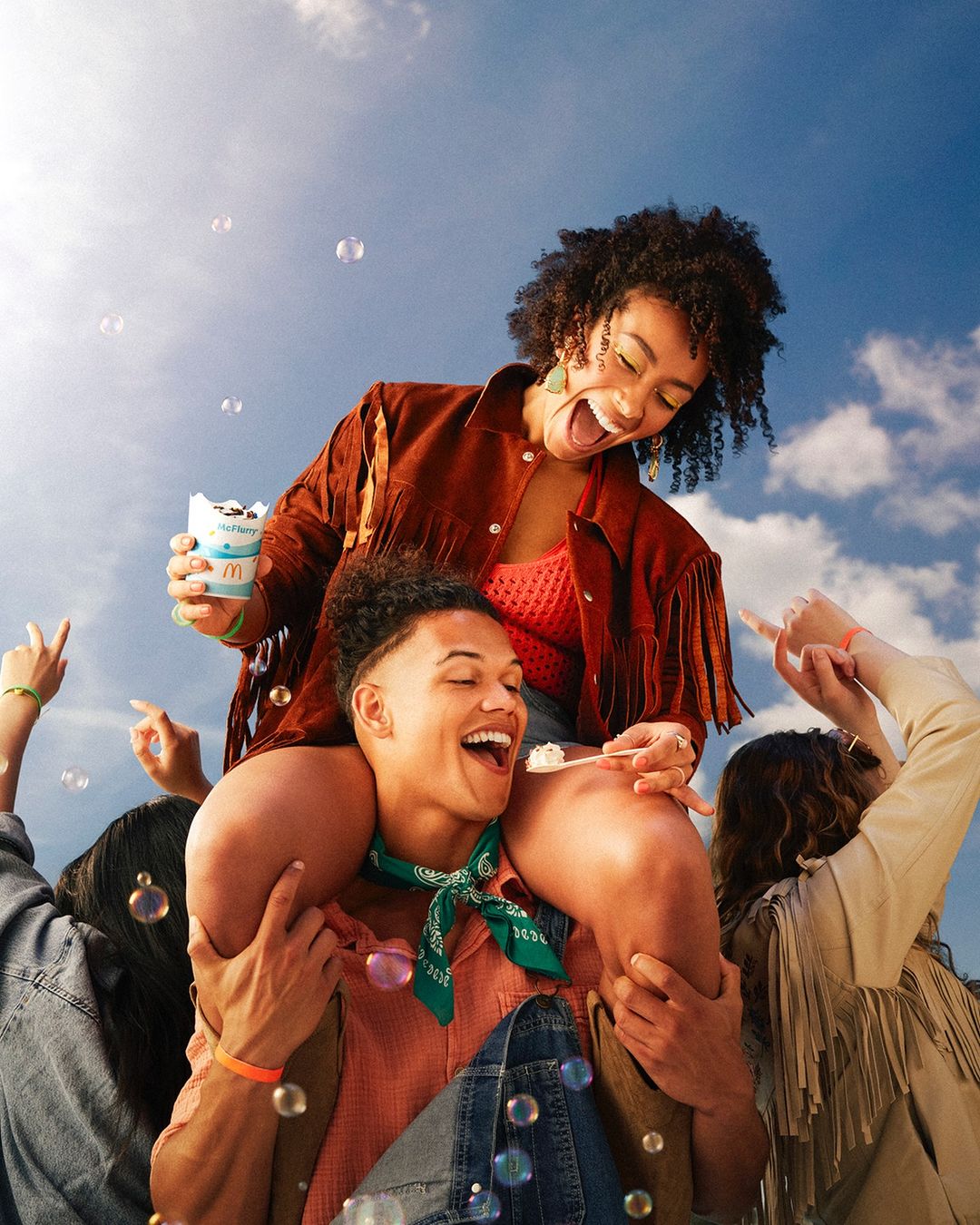 Young woman enjoys a sweet treat on a man's shoulders, surrounded by friends and soap bubbles against a cloudy sky.