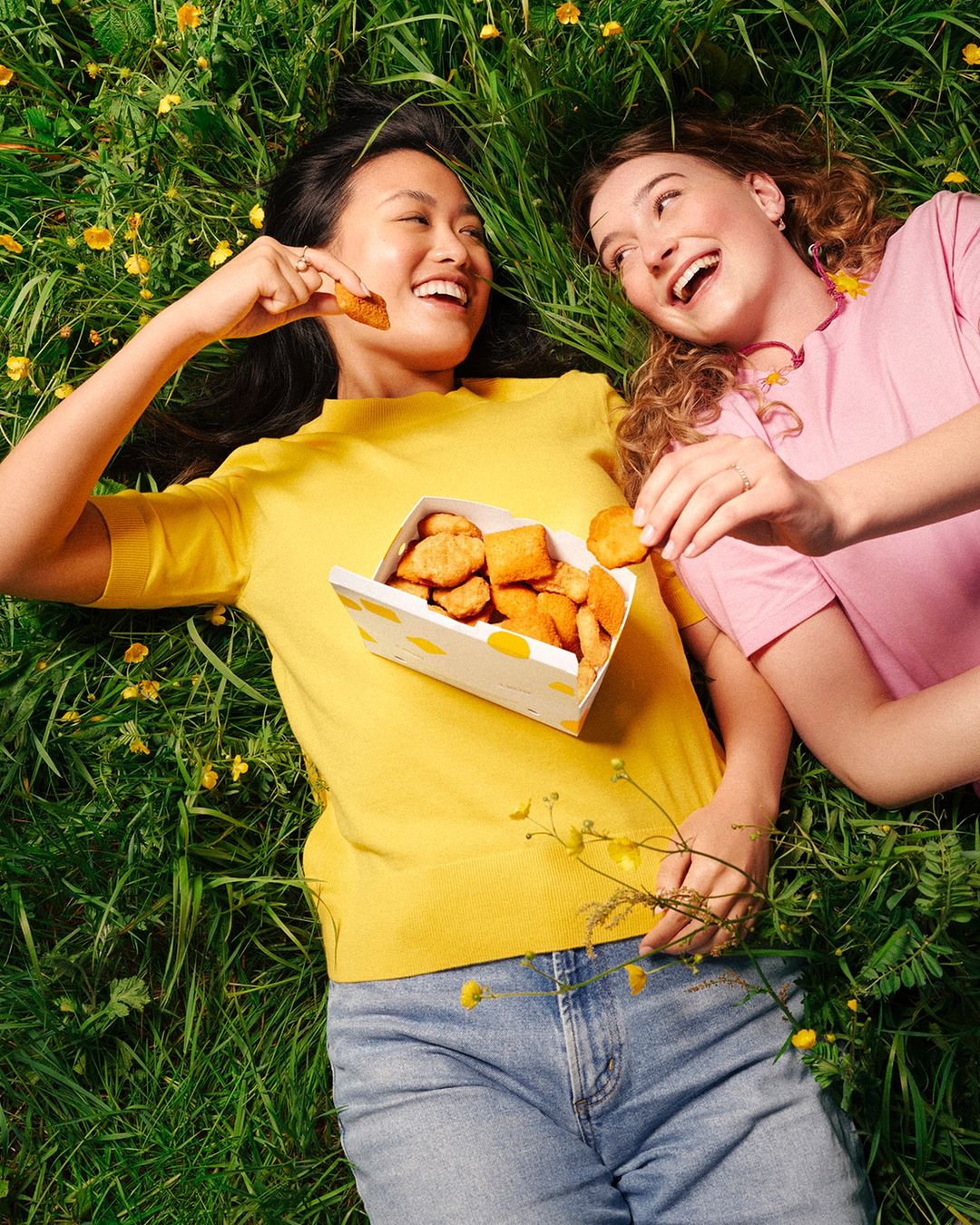 Two smiling women lying in a field of green grass and yellow flowers, holding a box of chicken nuggets and eating them.