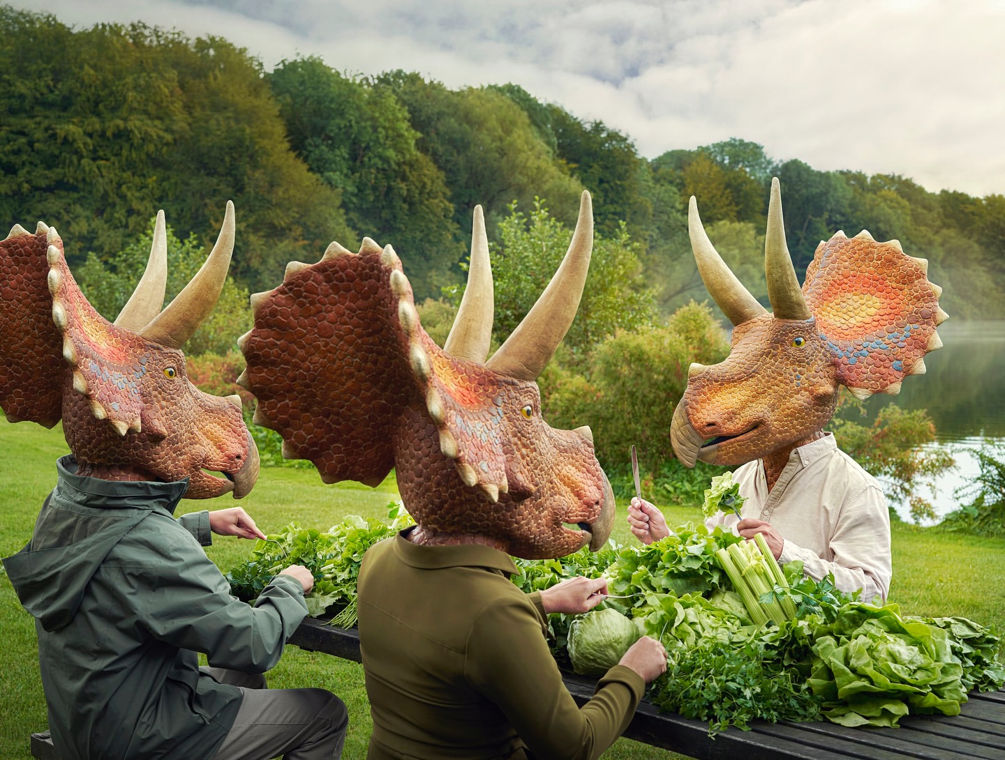 Three people wearing dinosaur head masks preparing fresh greens at a table in a green outdoor setting with a lake in the background.