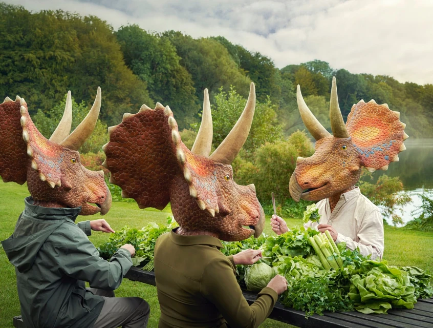 Three people wearing dinosaur head masks preparing fresh greens at a table in a green outdoor setting with a lake in the background.