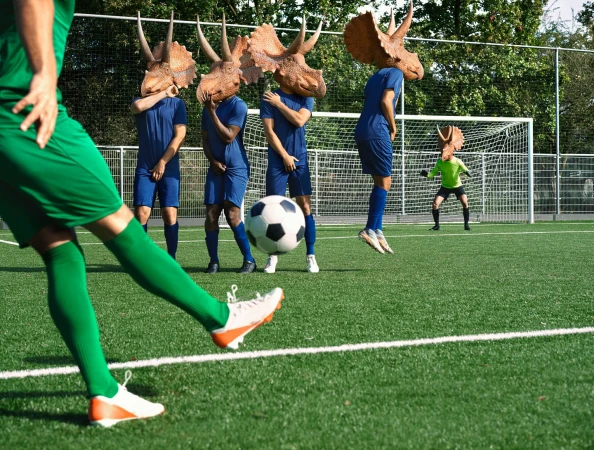 Soccer team with triceratops masks prepares for a free kick on a green soccer field on a sunny day. One player kicks the ball.