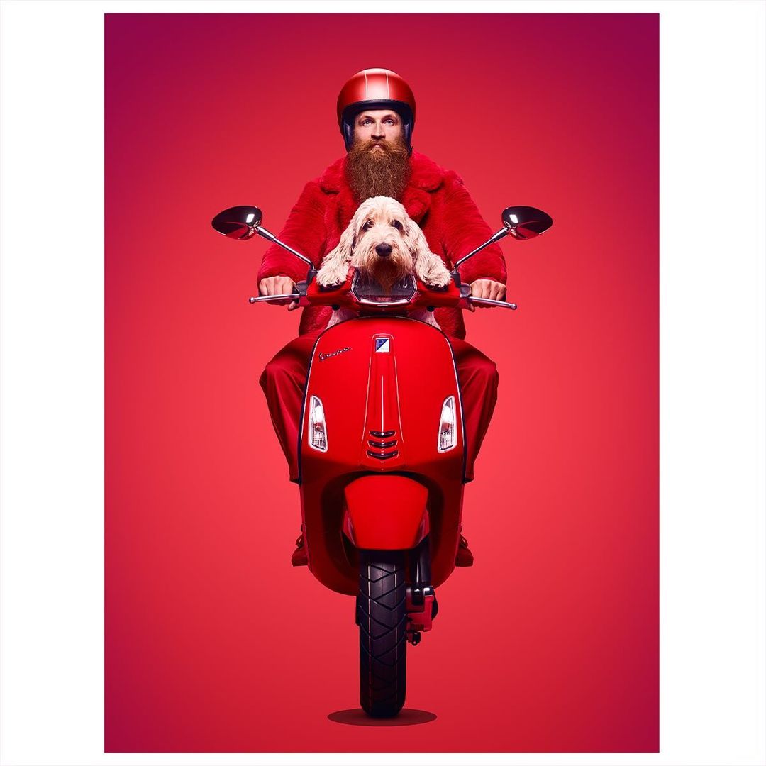 Adult male with a beard and a helmet riding a red motor scooter with a dog sitting in front of him against a red background.