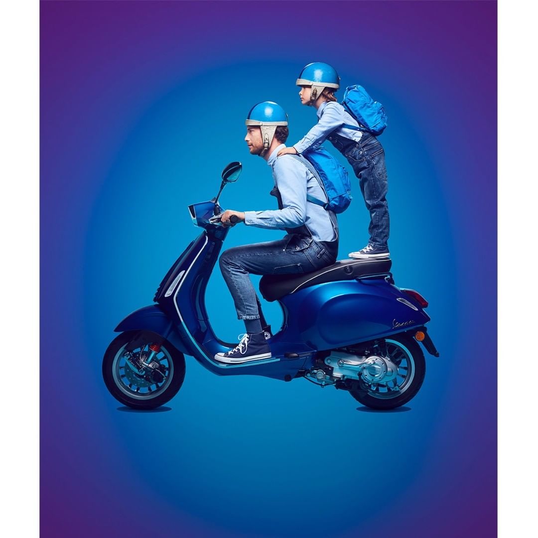 Father and child on blue scooter with matching blue helmets and backpacks against blue gradient background in a studio setting.