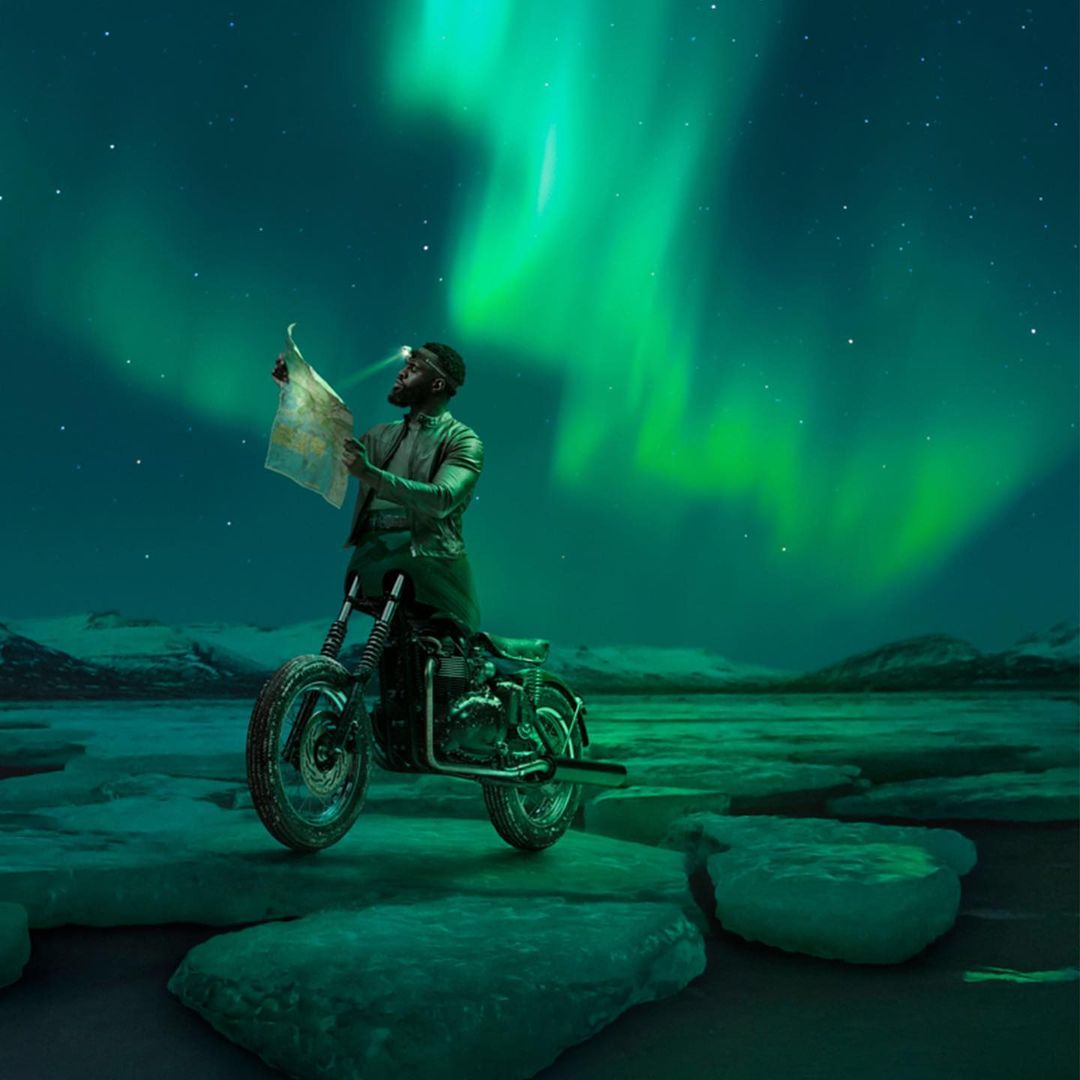 Man on motorcycle navigates icy terrain with map under aurora borealis. Adventure travel scene lit with headlamp under night sky.