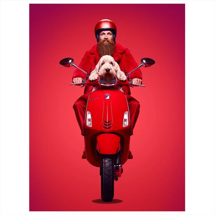 Bearded man in red faux fur coat and helmet riding a red scooter with a small dog