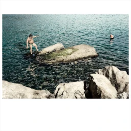 Man in swimming trunks wearing goggles stepping onto coastal rock formation in water by Jaap Vliegenthart