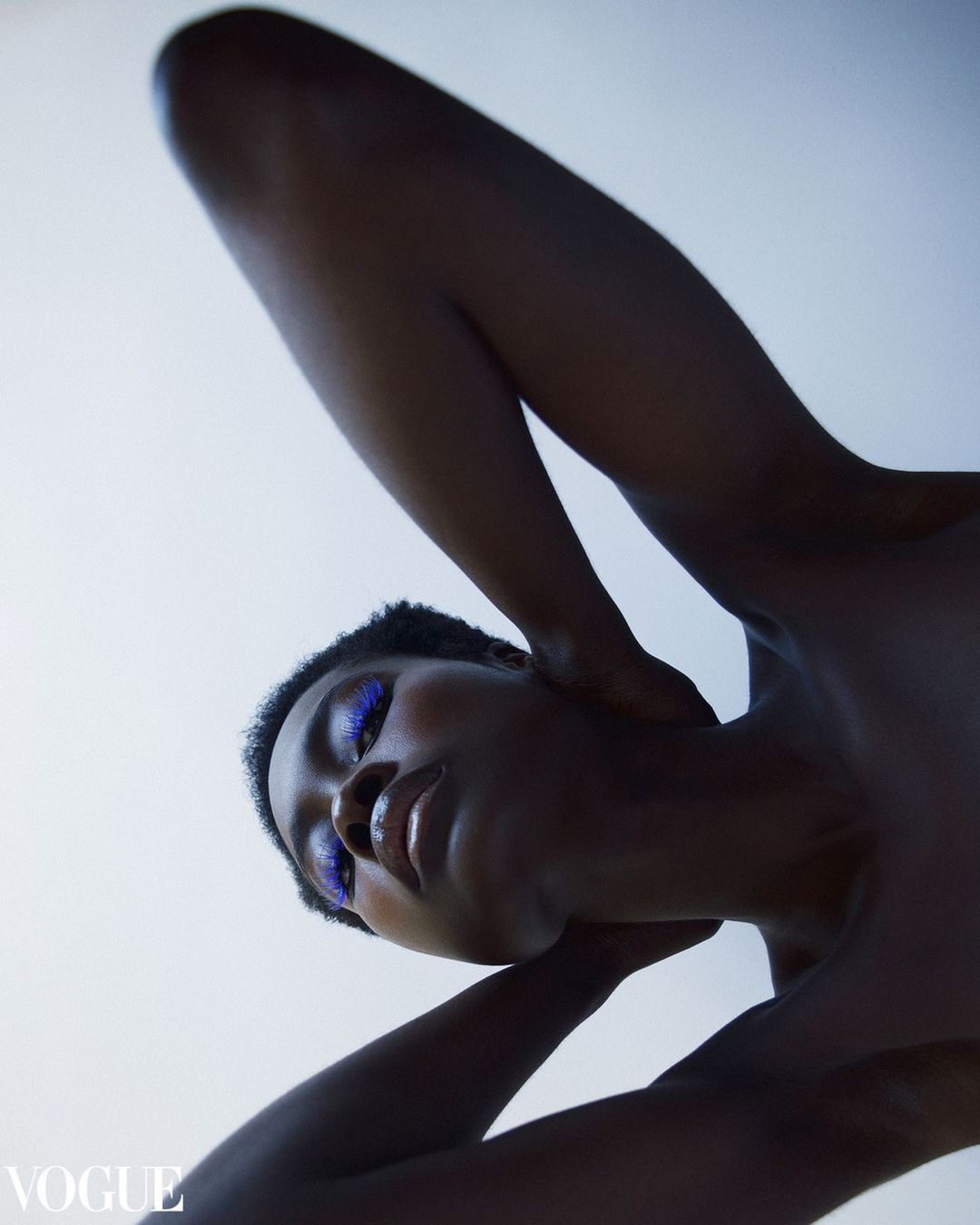 A dark-skinned woman with bright purple eyeshadow poses in a low-angle shot, illuminated against a white background, showcasing beauty.