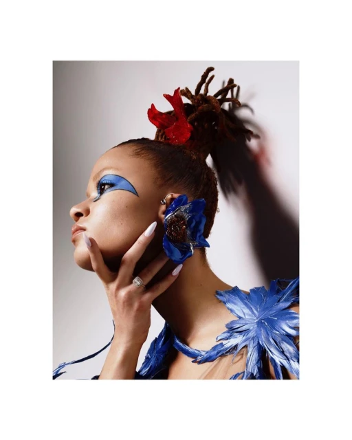 Close-up studio shot of a woman with dreadlocks, creative blue makeup, and blue feather-like decorations, set against a gradient background.