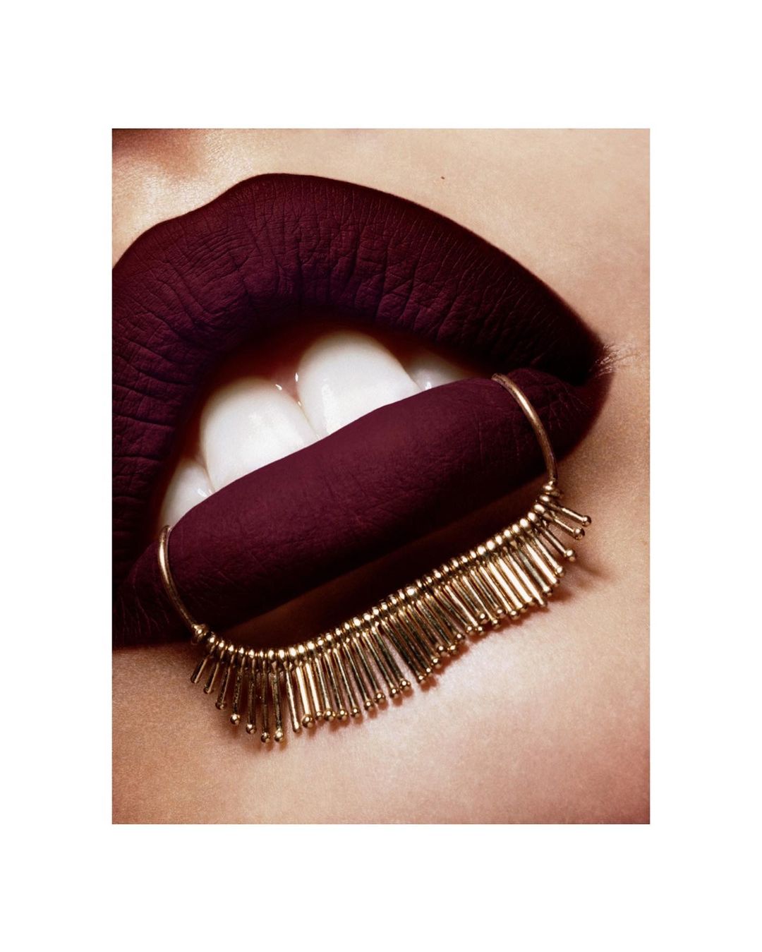 Close-up studio shot of a woman's lips with dark burgundy lipstick and a gold beaded accessory attached to the lower lip.