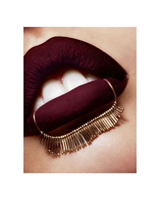 Close-up studio shot of a woman's lips with dark burgundy lipstick and a gold beaded accessory attached to the lower lip.