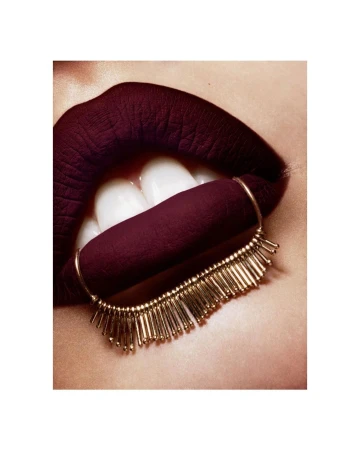 Close-up studio shot of a woman's lips with dark burgundy lipstick and a gold beaded accessory attached to the lower lip.