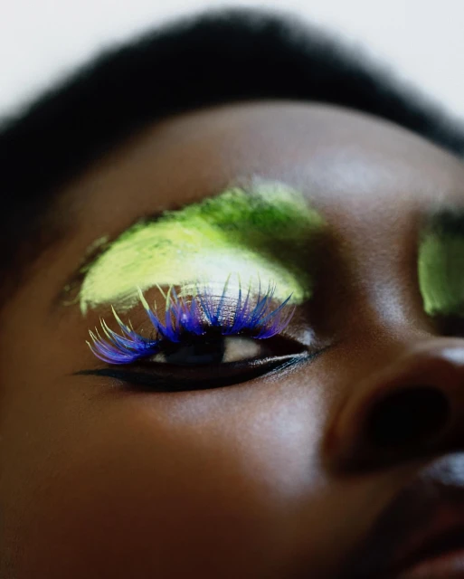 Close-up portrait of an adult female with dark skin, dramatic eye makeup with blue false eyelashes and green and yellow eyeshadow.