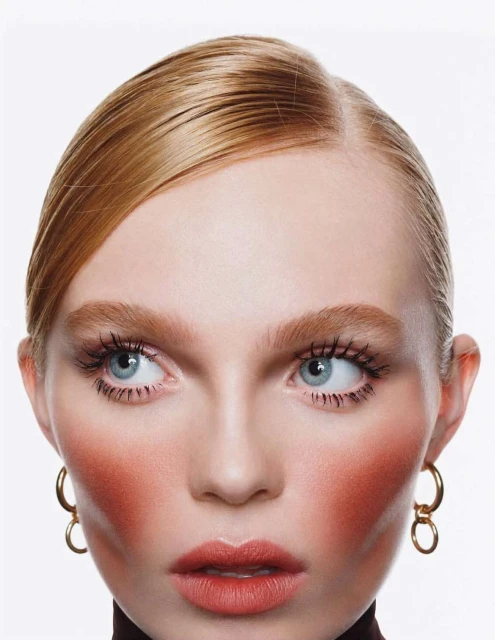 Close-up portrait of a fair-skinned woman with blonde hair, wearing gold hoop earrings, with bright makeup and a brown turtleneck top.