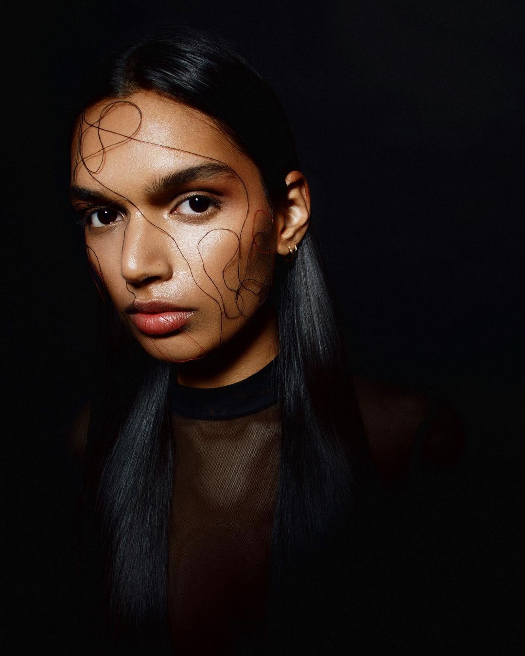 Close-up portrait of an adult woman with dark hair, fair skin, and light makeup. Black geometric designs on her face against a black background.