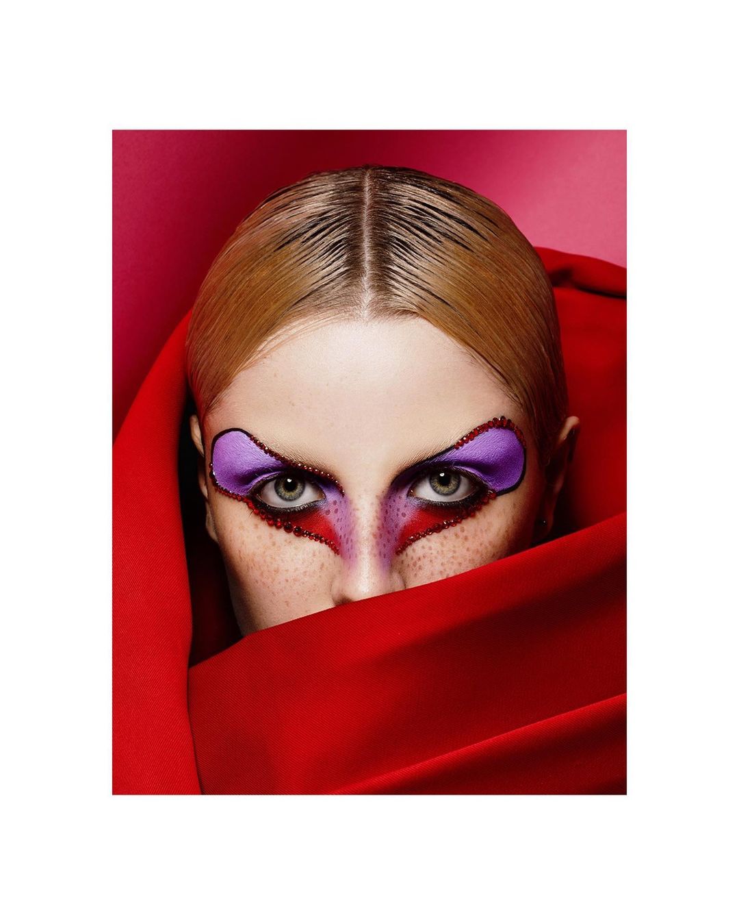 Close-up studio portrait of a blonde female model with dramatic purple and red makeup partially covered by a draped red cloth.