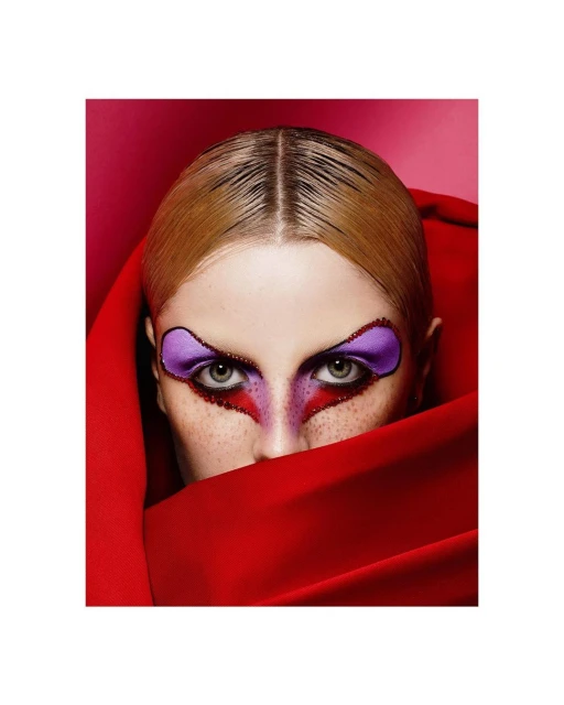 Close-up studio portrait of a blonde female model with dramatic purple and red makeup partially covered by a draped red cloth.