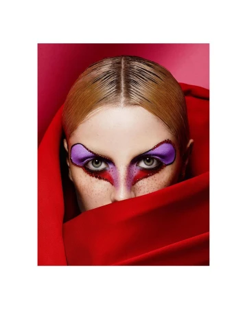 Close-up studio portrait of a blonde female model with dramatic purple and red makeup partially covered by a draped red cloth.