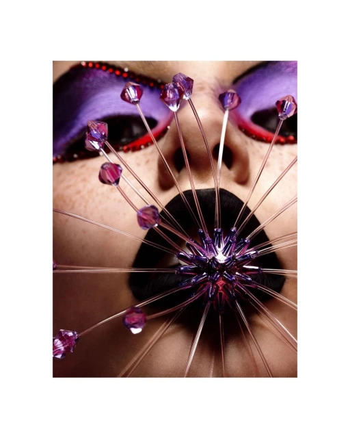 Eye-level close-up of a human face with purple makeup and an elaborate face decoration with bead accents and thin metallic spikes.