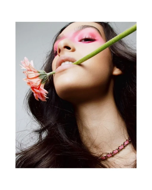 Close-up of a woman with bright pink makeup on her eyes holding a pink gerbera daisy in her mouth against a light gray background.