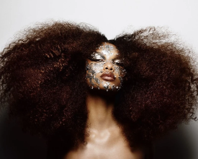 Close-up portrait of a black woman with large afro hairstyle and foil makeup, against a plain white backdrop in a studio setting.