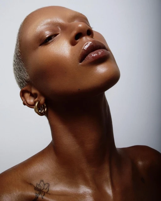 Close-up studio portrait of a Black woman with a shaved head, shiny skin, glossy lips, gold hoop earrings, and a floral tattoo.