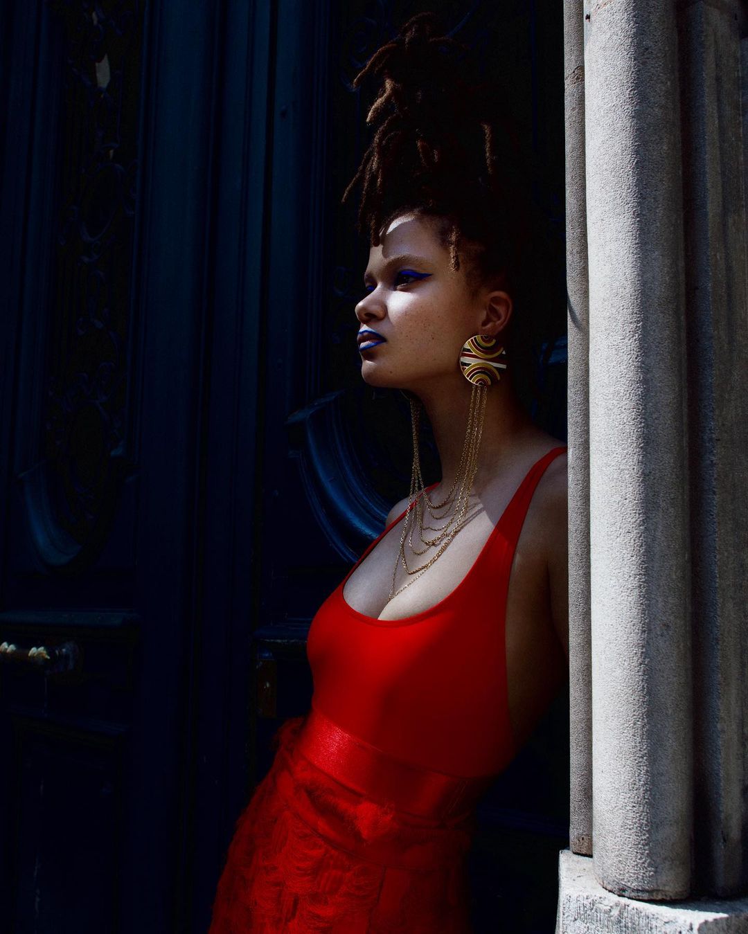 Portrait of an adult female wearing a red dress and large earrings, posing next to a stone pillar with dark blue background.