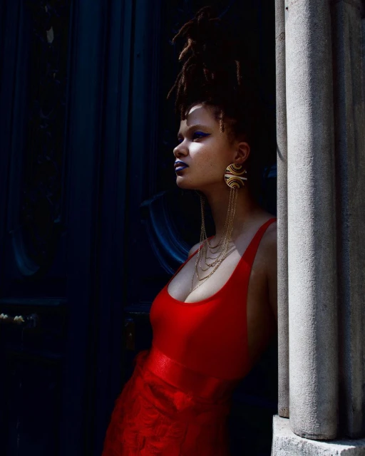 Portrait of an adult female wearing a red dress and large earrings, posing next to a stone pillar with dark blue background. 