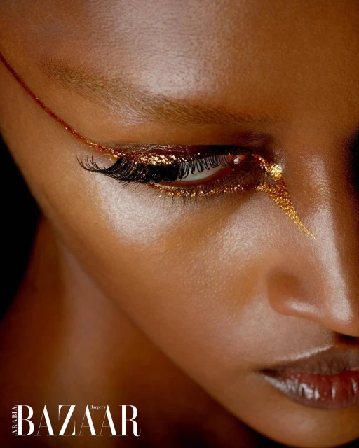 Close-up portrait of a Black woman with golden glitter eyeliner and sleek dark skin, posing in a studio with dark background.