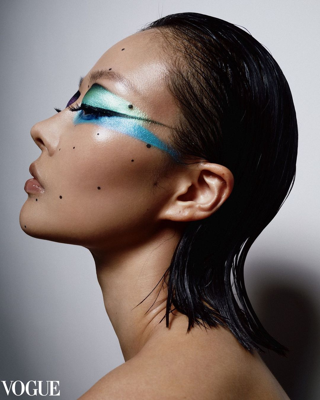 Profile shot of an Asian woman with colorful eye makeup and wet-look hair against a neutral backdrop. Vogue logo is visible.