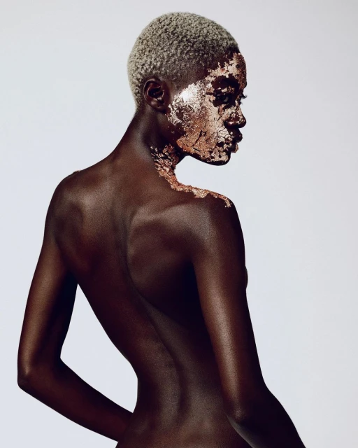 Close-up studio shot of a bald, dark-skinned woman with gold foil accents on her face and shoulder, looking over her shoulder at the camera.