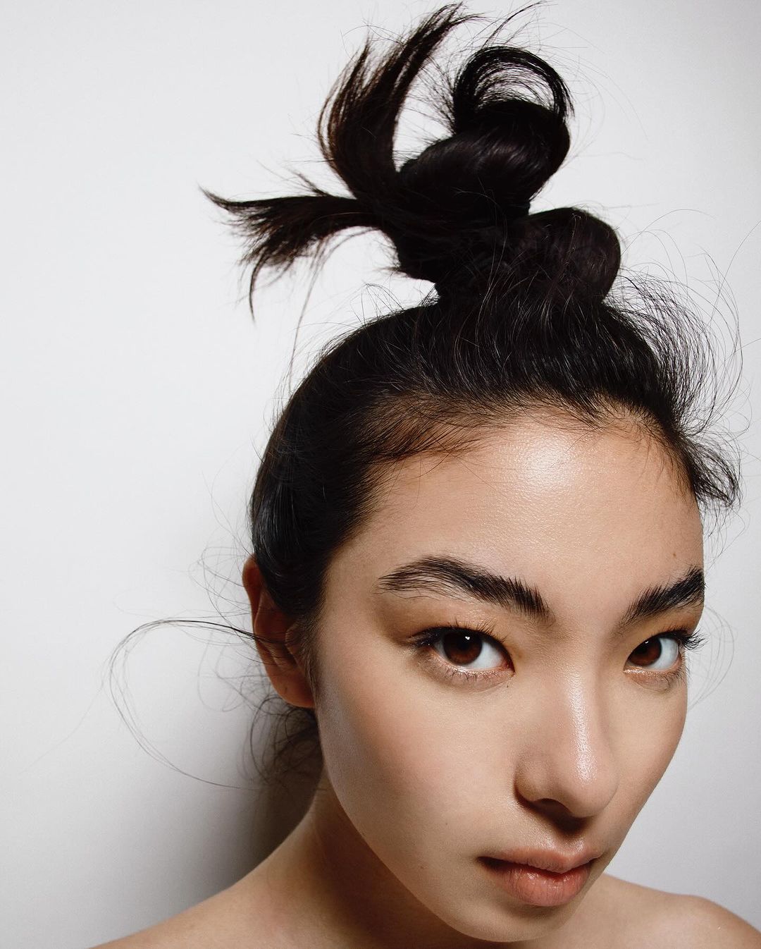 Headshot of a woman with dark hair styled in a high bun, light complexion, light makeup, fair skin, and neutral expression on a white background.