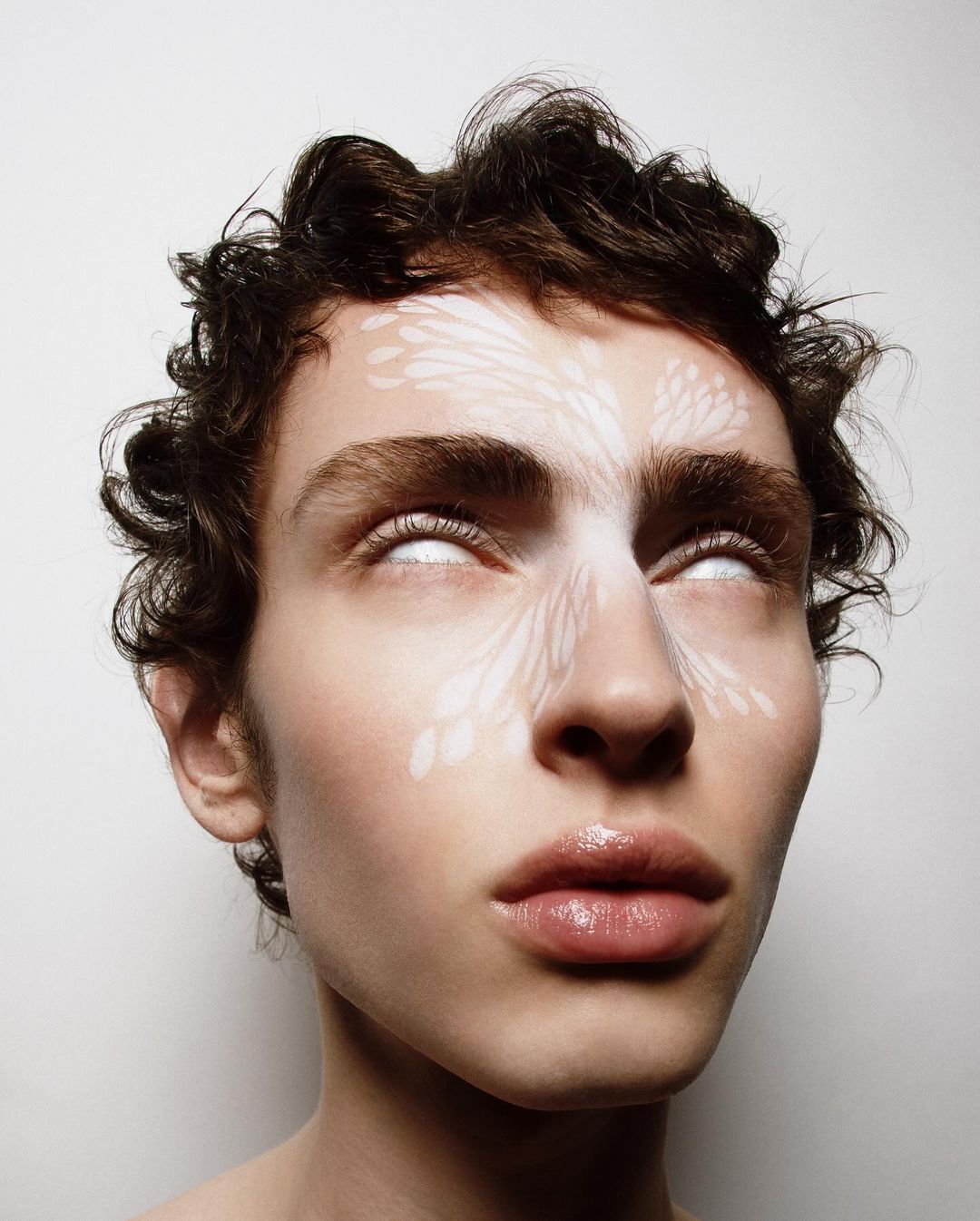 Close-up of a young adult with light skin, curly dark hair and white patterned face paint, staring upwards with white, opaque eyes.