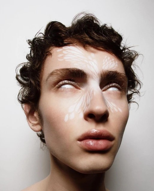 Close-up of a young adult with light skin, curly dark hair and white patterned face paint, staring upwards with white, opaque eyes.