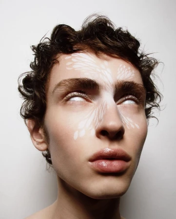 Close-up of a young adult with light skin, curly dark hair and white patterned face paint, staring upwards with white, opaque eyes.