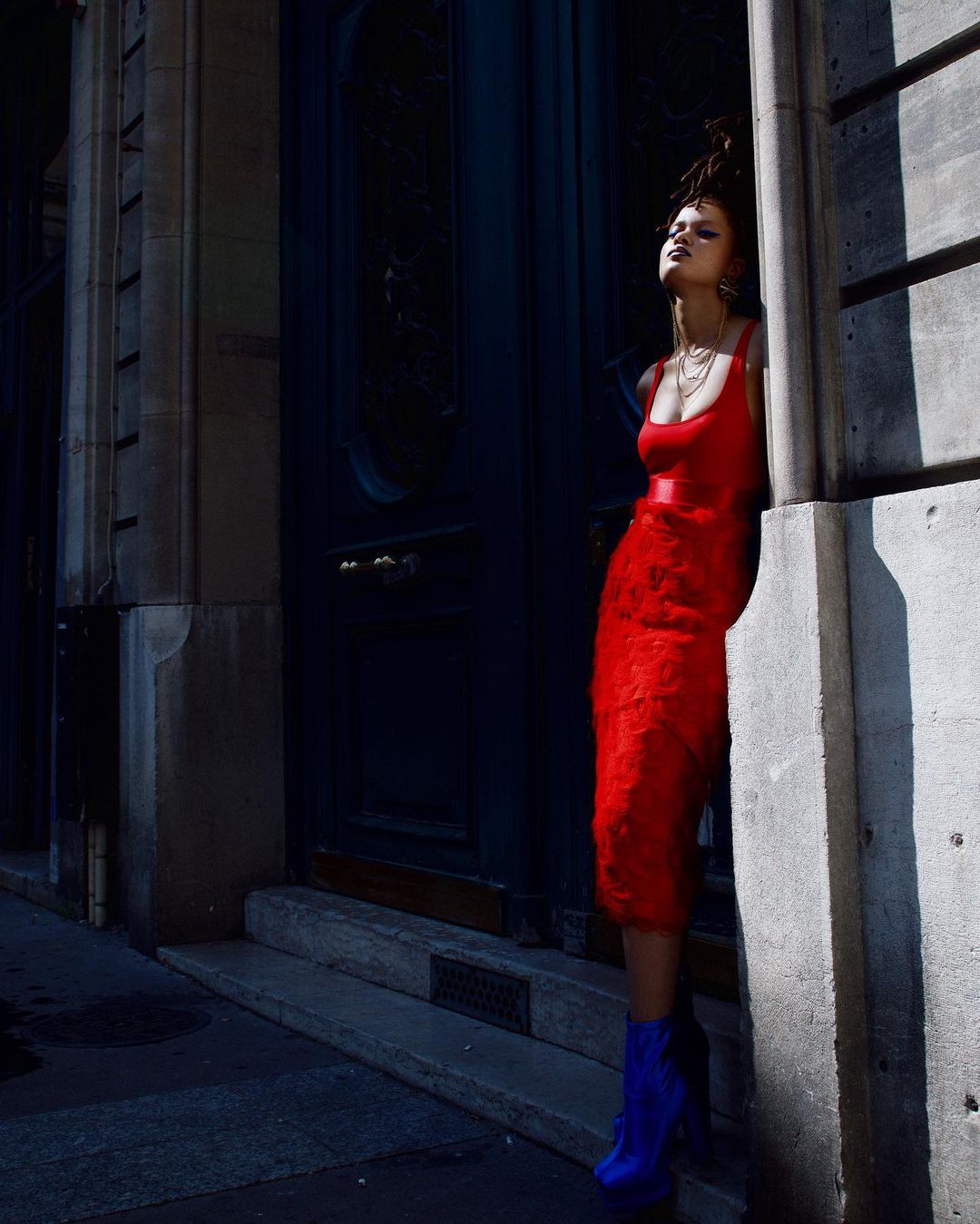 Fashion shot of a woman in a red dress posing against a building in an urban setting with bright sunlight.