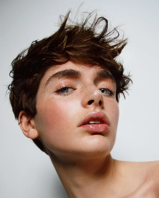 Close-up portrait of a young adult woman with brown hair and light eyes, fair skin, freckles, glossy lips, and light makeup against a white background.