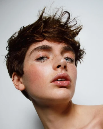 Close-up portrait of a young adult woman with brown hair and light eyes, fair skin, freckles, glossy lips, and light makeup against a white background.