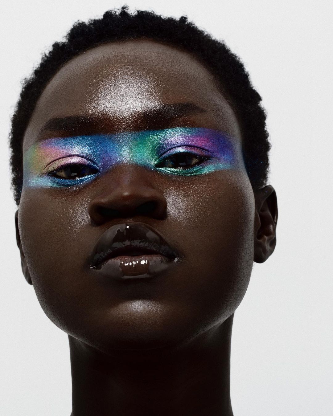Close-up portrait of a dark-skinned female with iridescent blue, green, and purple makeup across her eyebrows, glossy lips, and slicked hair.