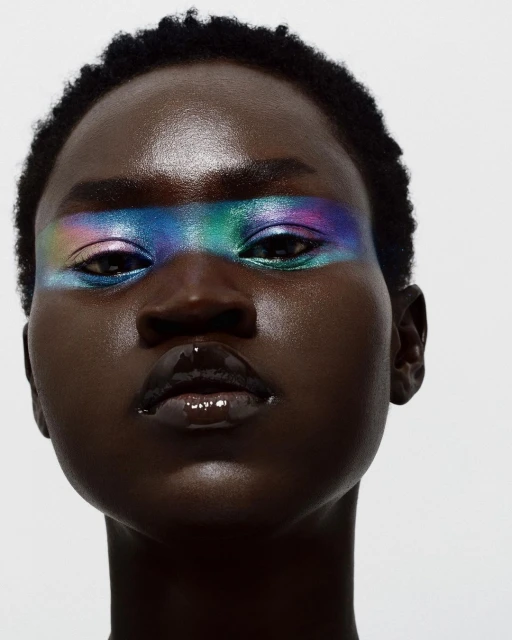 Close-up portrait of a dark-skinned female with iridescent blue, green, and purple makeup across her eyebrows, glossy lips, and slicked hair.